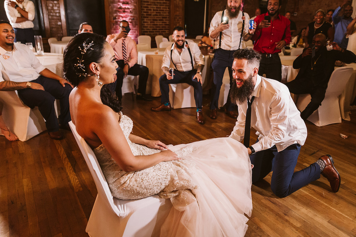 bride sits on a white chair. groom is on his knees on a wood floor in front of her as he prepares to take off her garter