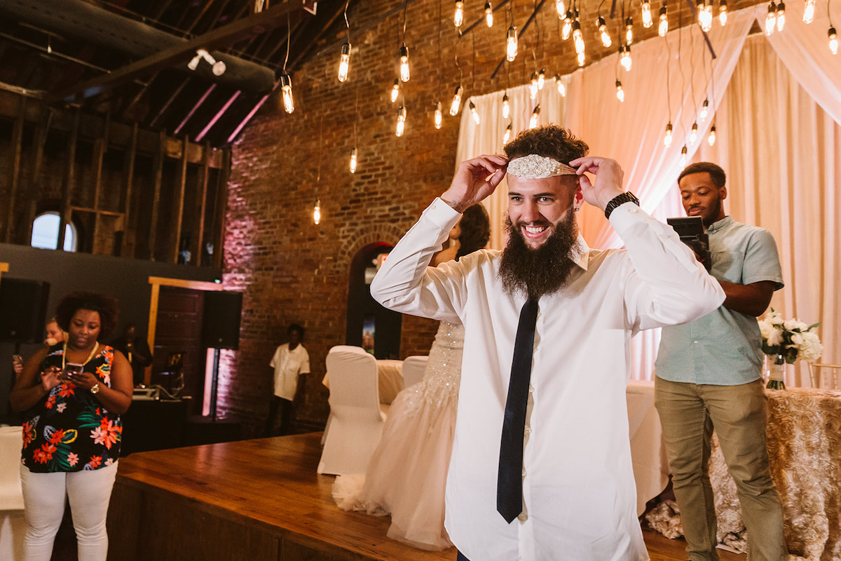 groom places bride's white lacy garter around his head under open rafters of The Church on Main in Chattanooga