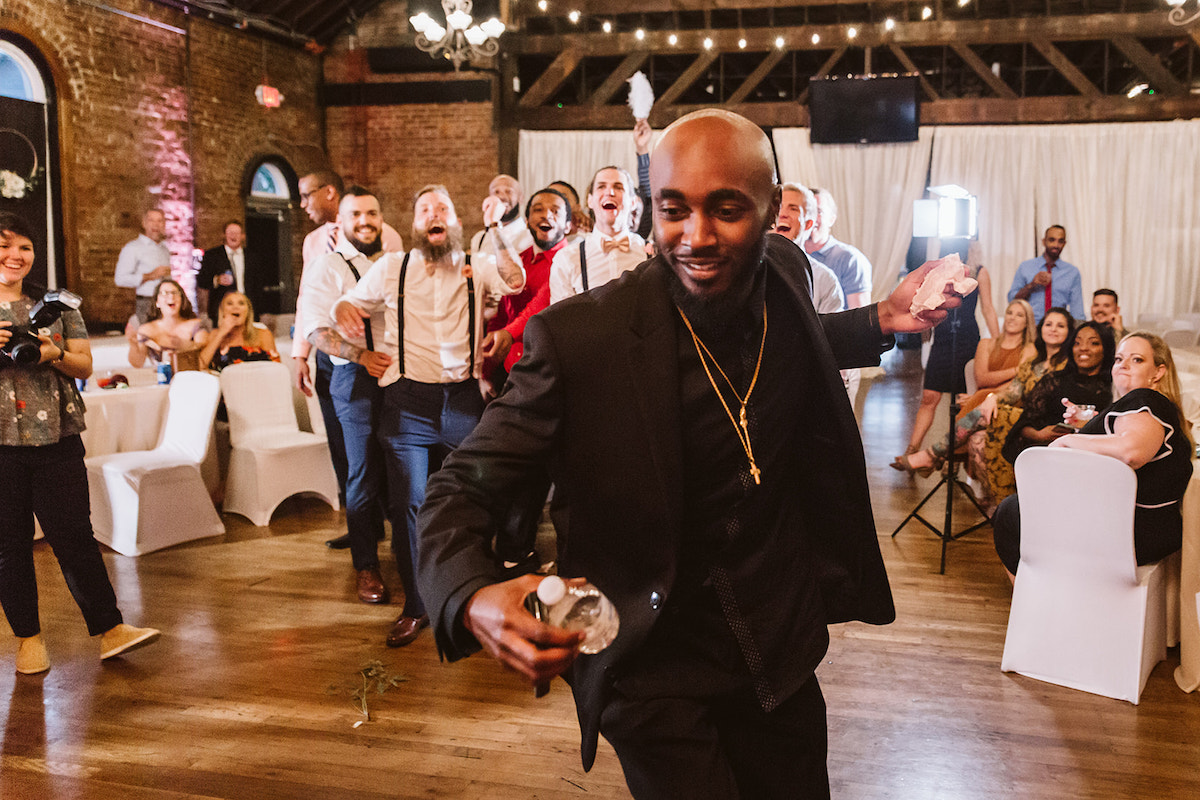 Man in black suit and shirt dances while holding the bride's garter as other men watch