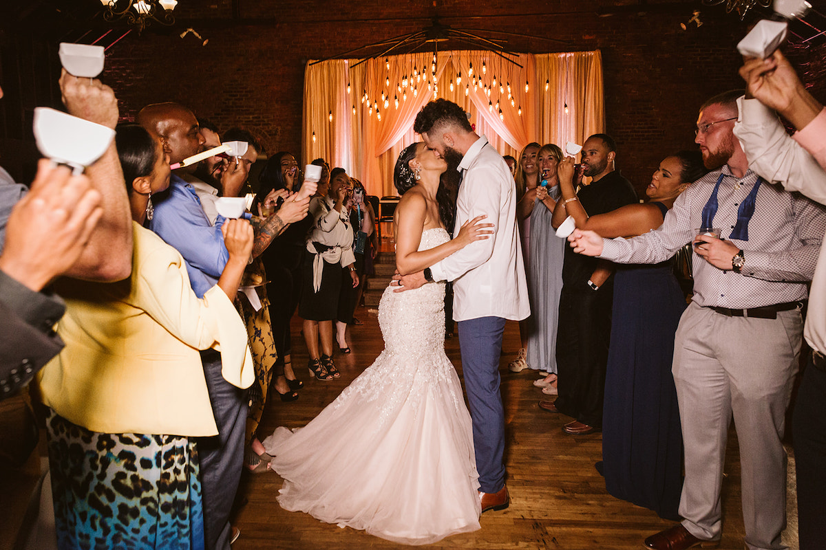 Bride and groom kiss while guests clap and ring white bells around them