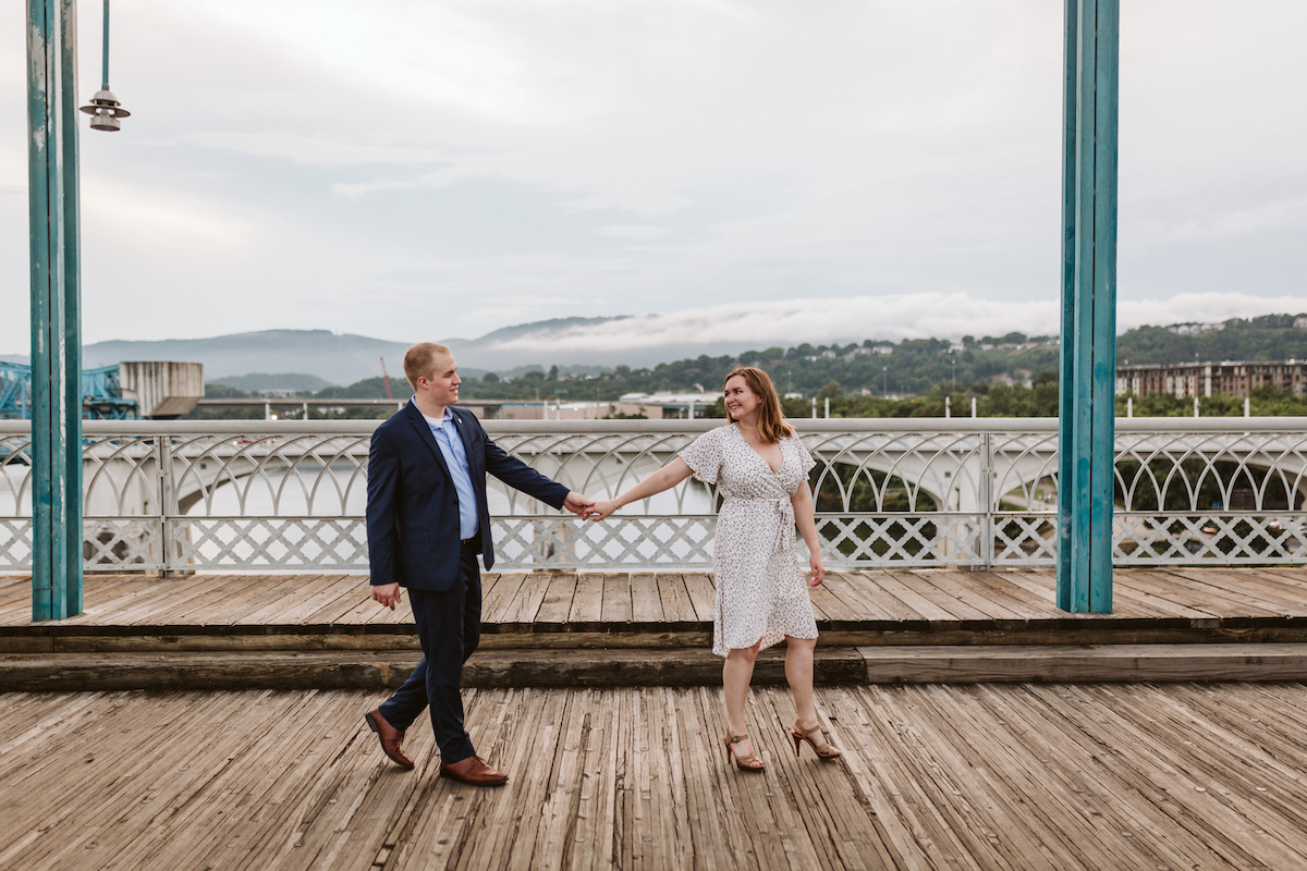 man in blue suit and brown shoes and woman in wrap-around knee-length dress hold hands on a bridge for engagement photos