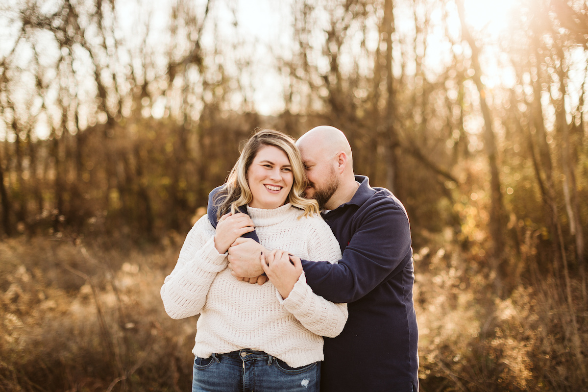 man in dark collared shirt cuddles over the shoulders of woman in cream turtleneck sweater during their engagement photos
