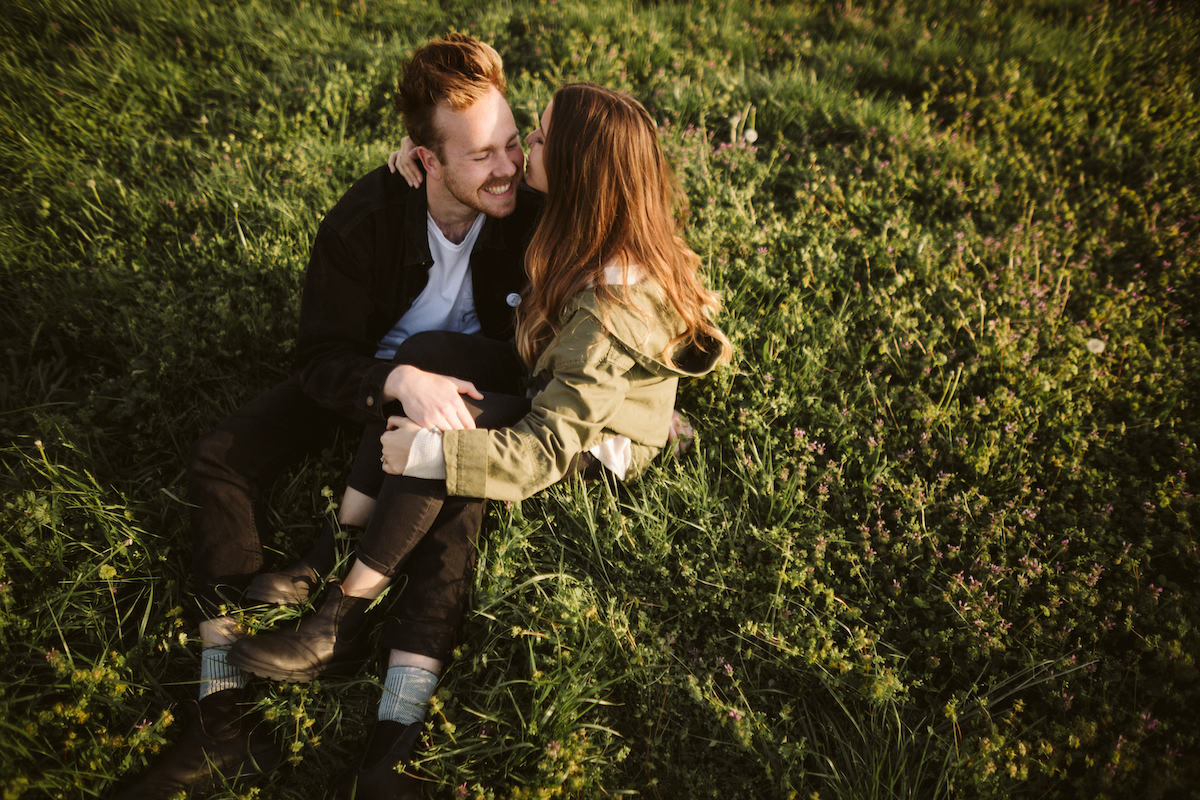 woman in black jeans and green jacket with her legs across mans knee as they sit in the grass during engagement photos