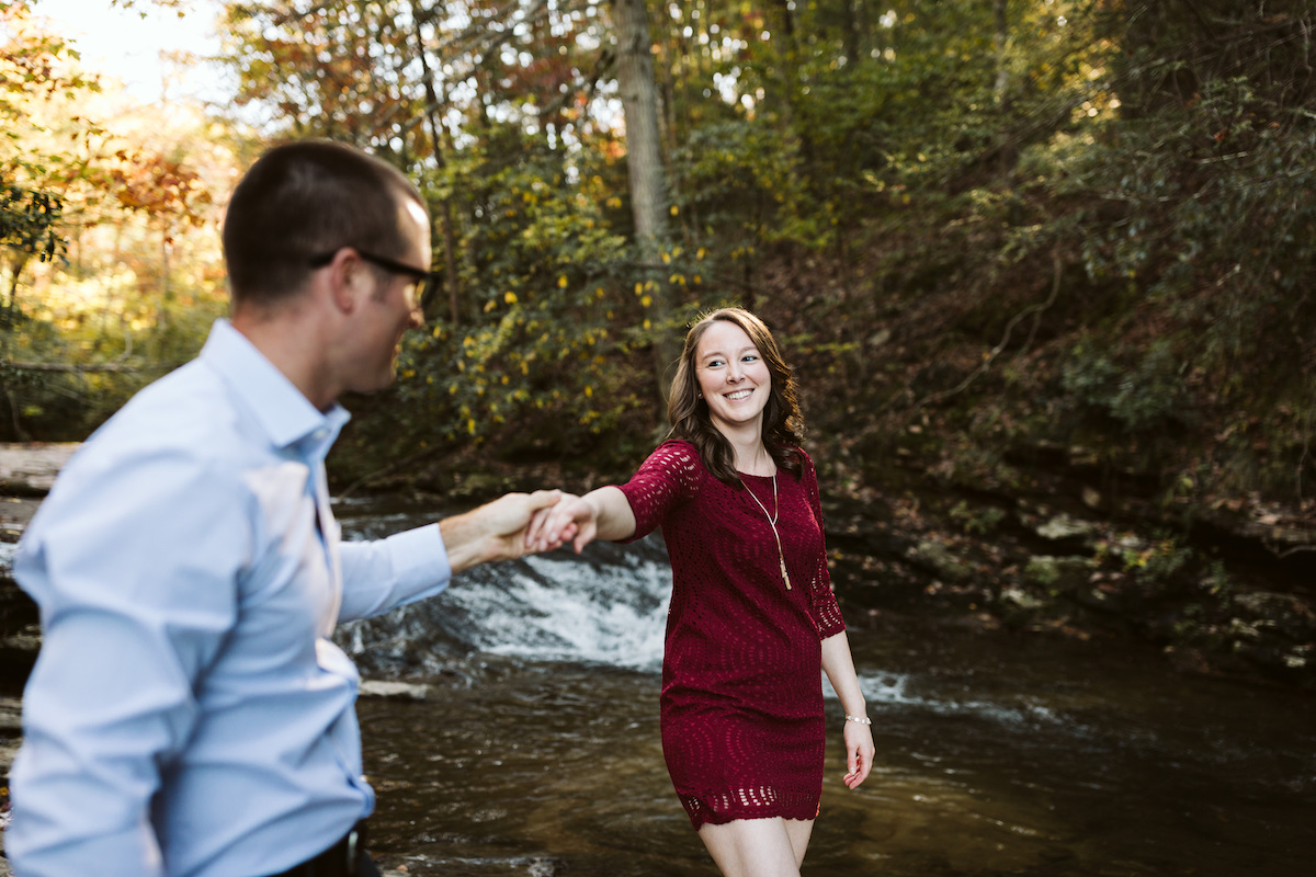 woman in short maroon dress holds hands with man in blue button down shirt next to a flowing creek