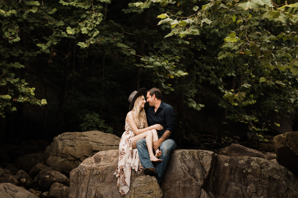 woman in floral sundress and black hat sits with man in blue jeans and black shirt on a large rock. her legs are in his lap.