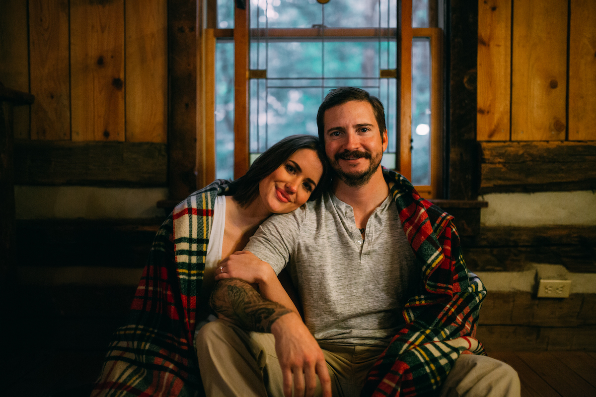 woman snuggles her head on man's shoulder as they sit beneath a shared plaid blanket in a wood cabin