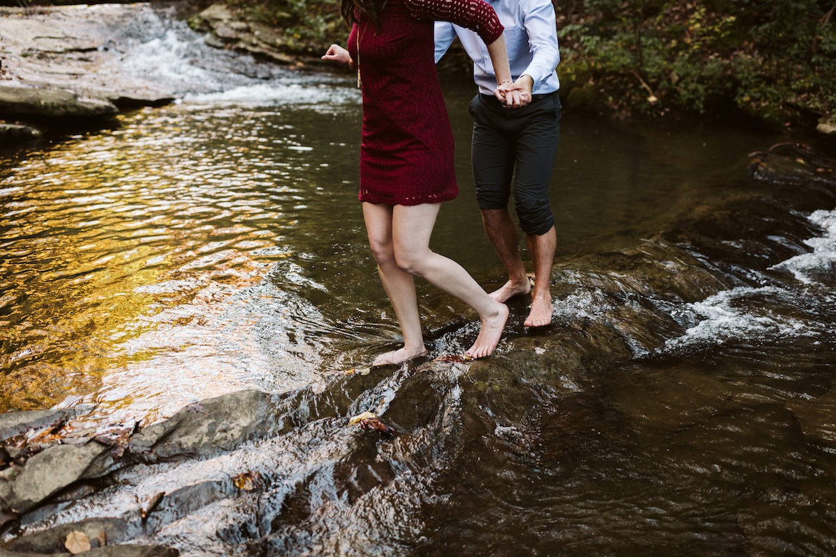 woman in short maroon dress and man with pants cuffed to his knees cross a rocky stream barefoot