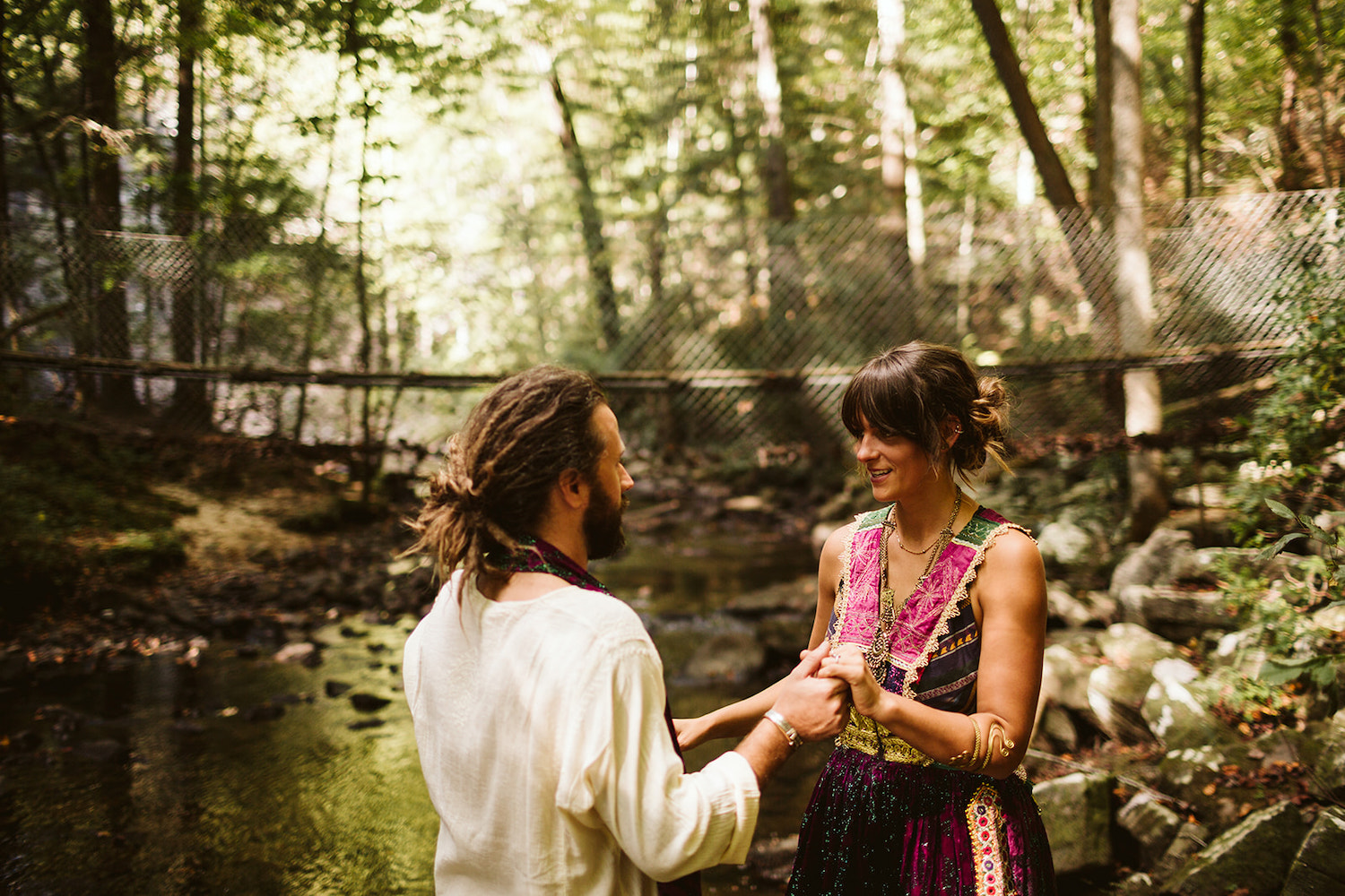 man and woman in colorful Bohemian style dress hold hands in the woods next to a creek