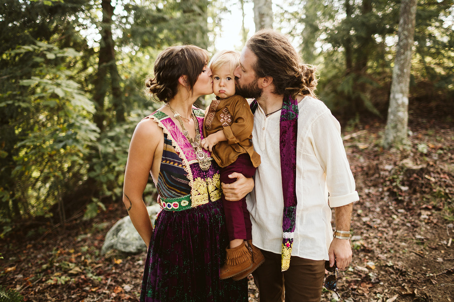 man and woman in colorful Bohemian dress hold a young blond child between them and kiss his cheeks