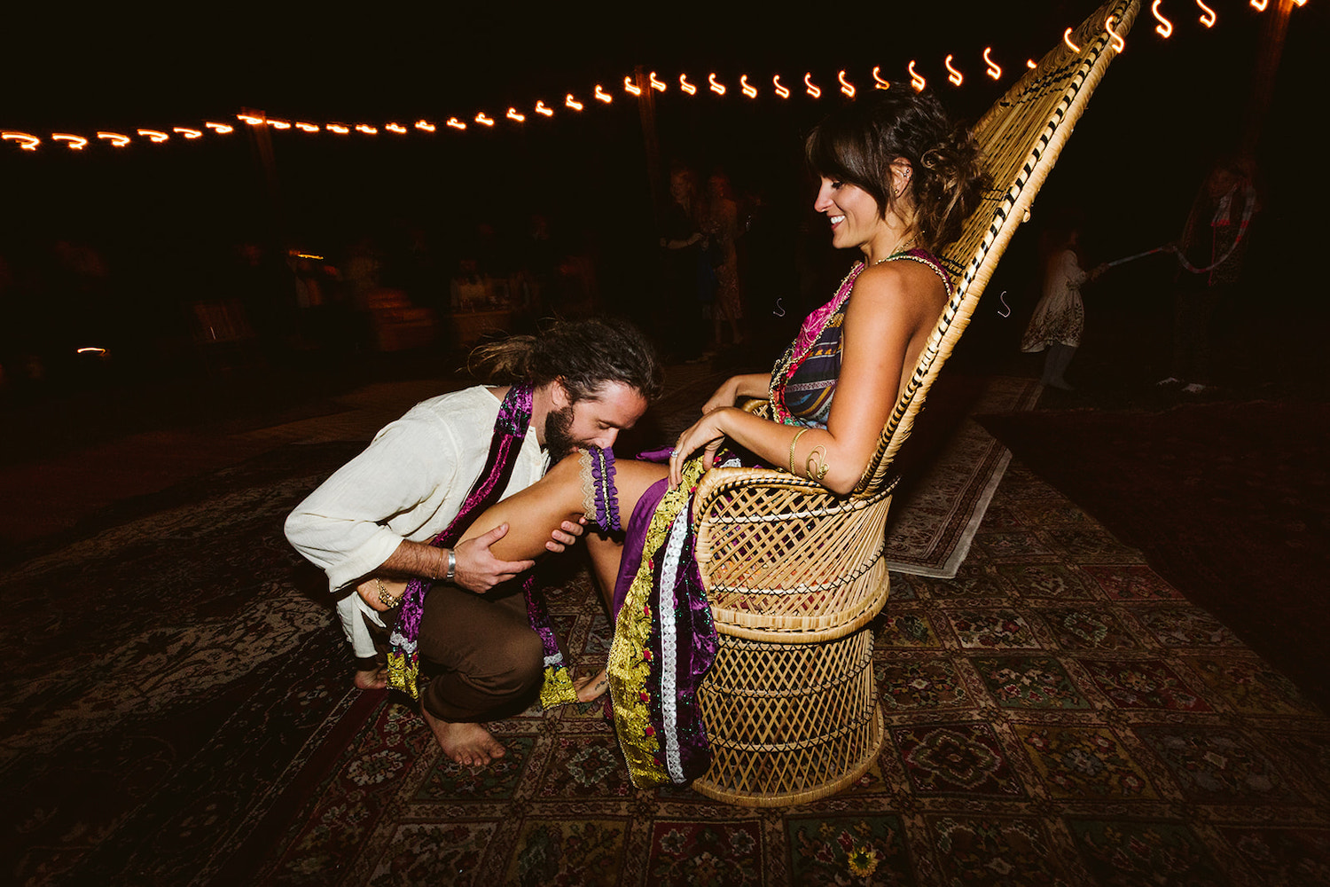 woman sits in tall wicker peacock chair while man removes her purple garter with his teeth