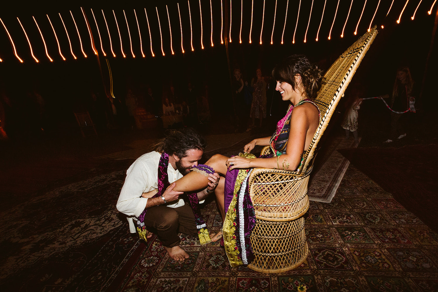 woman sits in tall wicker peacock chair while man removes her purple garter with his teeth