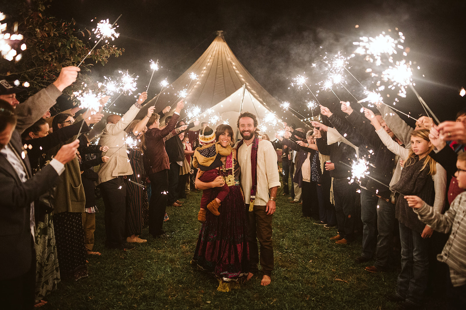 man and woman wearing bright gypsy dress walk under sparkler tunnel exit after their Hemlock Falls Bohemian festival wedding