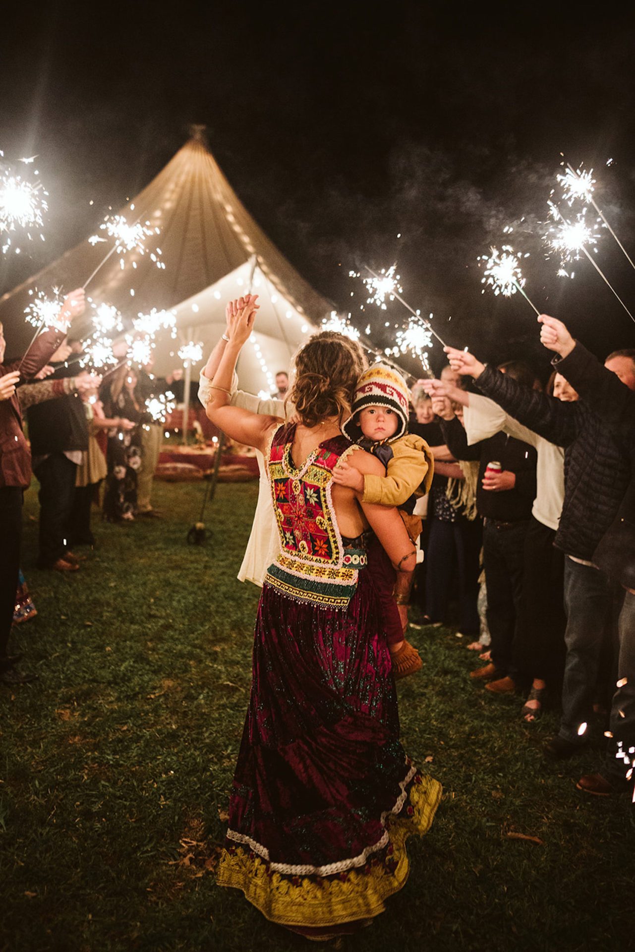 woman wearing bright Bohemian gypsy dress carries baby through sparkler tunnel exit after Chattanooga Hemlock Falls wedding