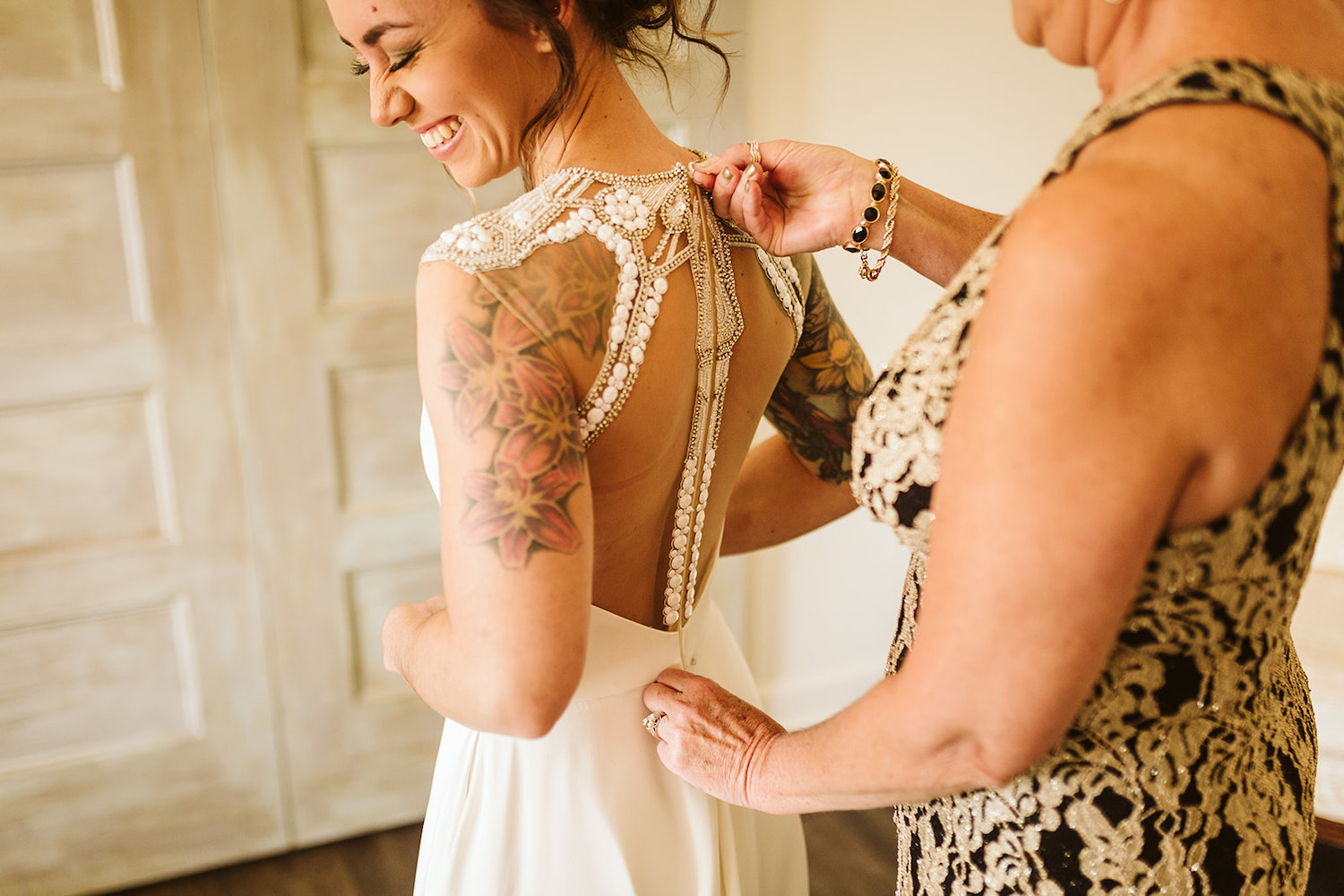 woman in lace dress zips the back of bride's gown while bride laughs