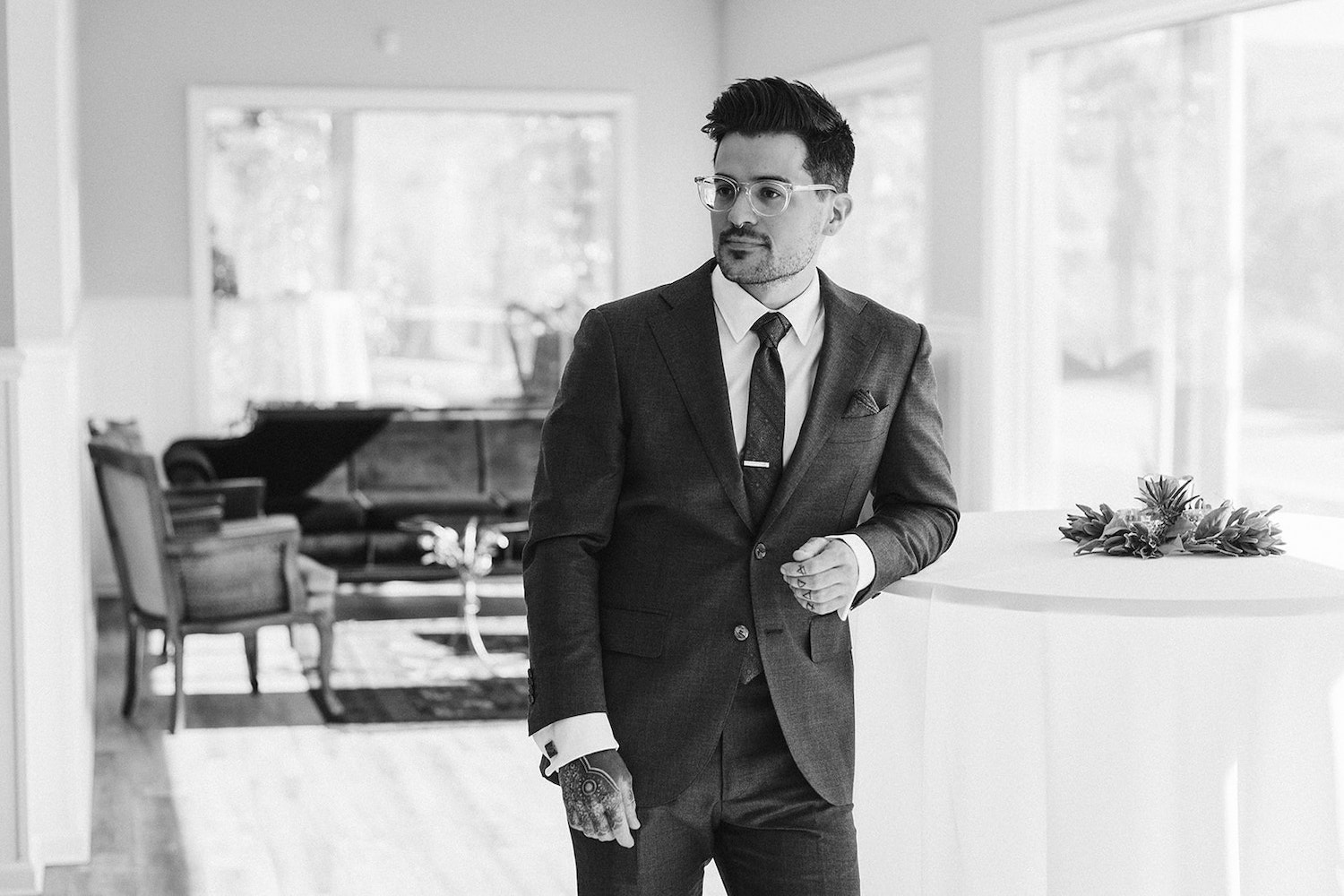 groom leans against a high table in front of large windows and vintage furniture