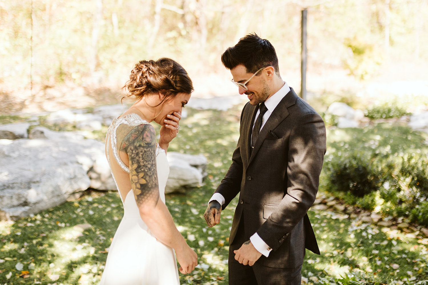 bride and groom laugh together in the shade of large trees as they share their first look