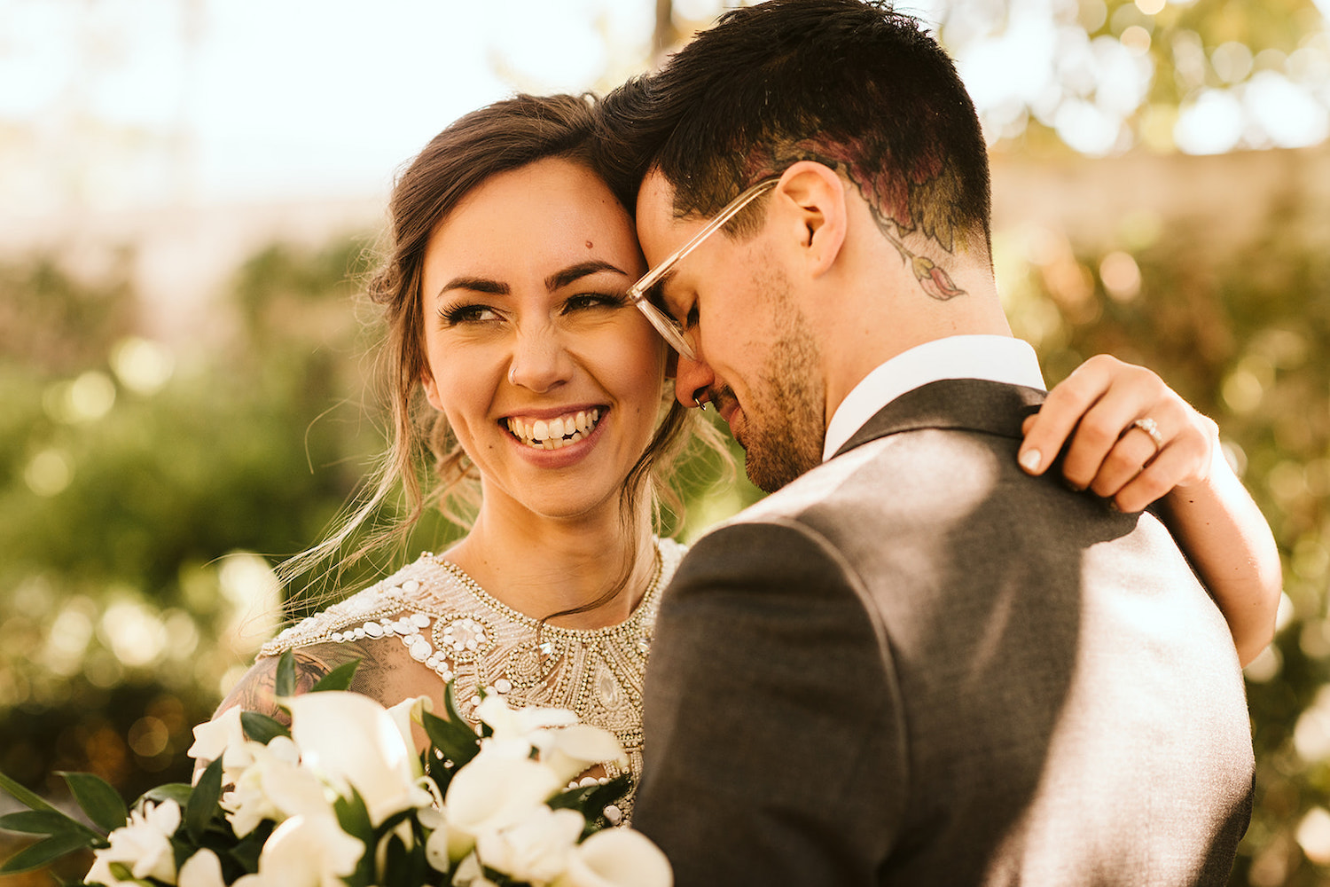 groom holds bride with his face on her temple. her arm is over his shoulder and she holds her large bouquet of white flowers