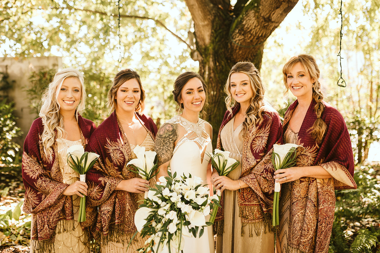 bride stands between bridesmaids who hold long stem calla lilies and wear maroon and gold scarves over their shoulders
