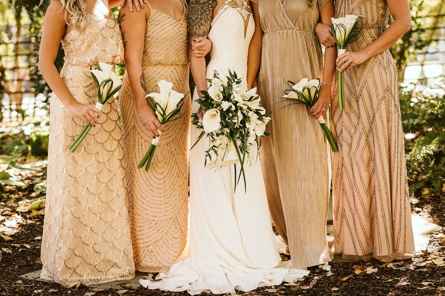 bride stands between bridesmaids who wear cream and gold dreses. they all hold white calla lilies.
