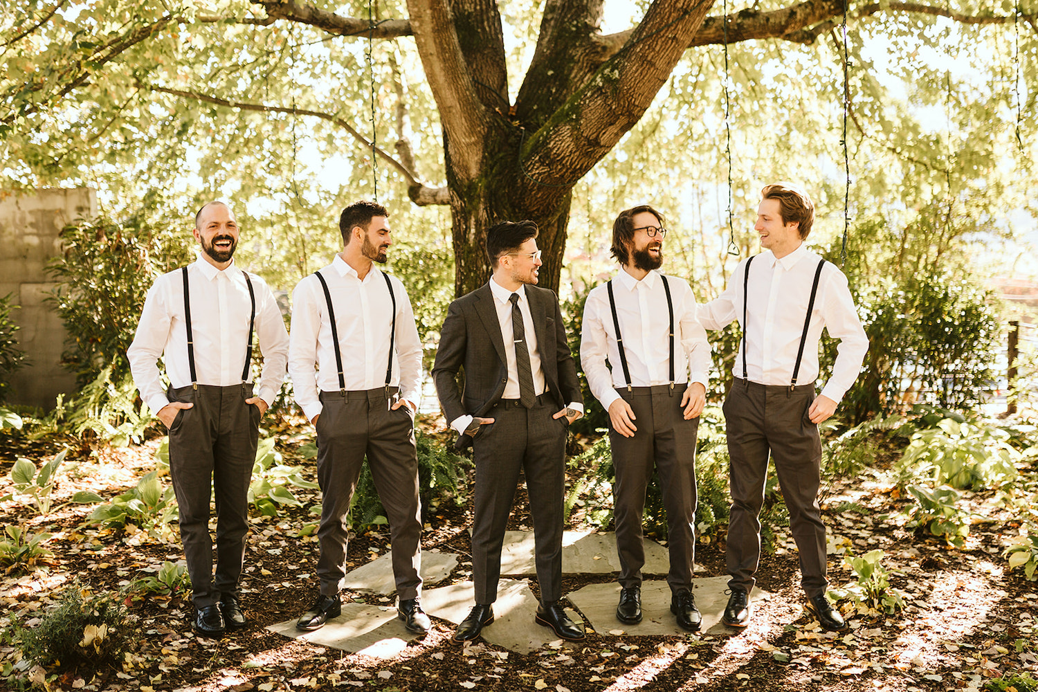 groom stands between groomsmen who wear dark pants, white shirts, and dark suspenders