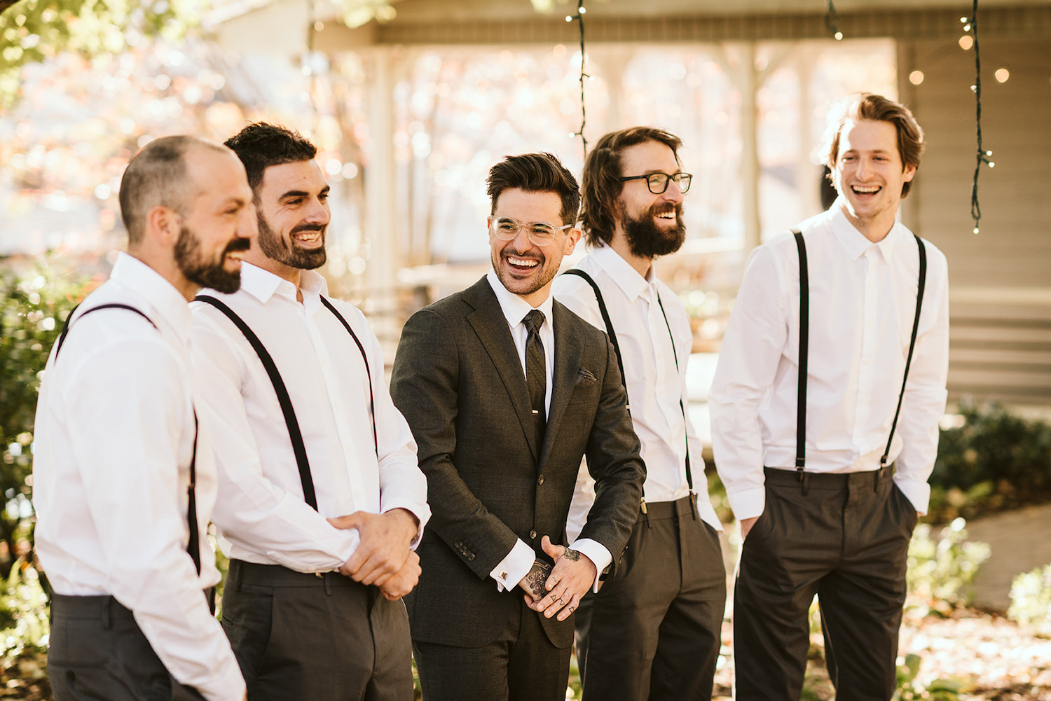 groom stands between groomsmen who wear dark pants, white shirts, and dark suspenders while they all laugh