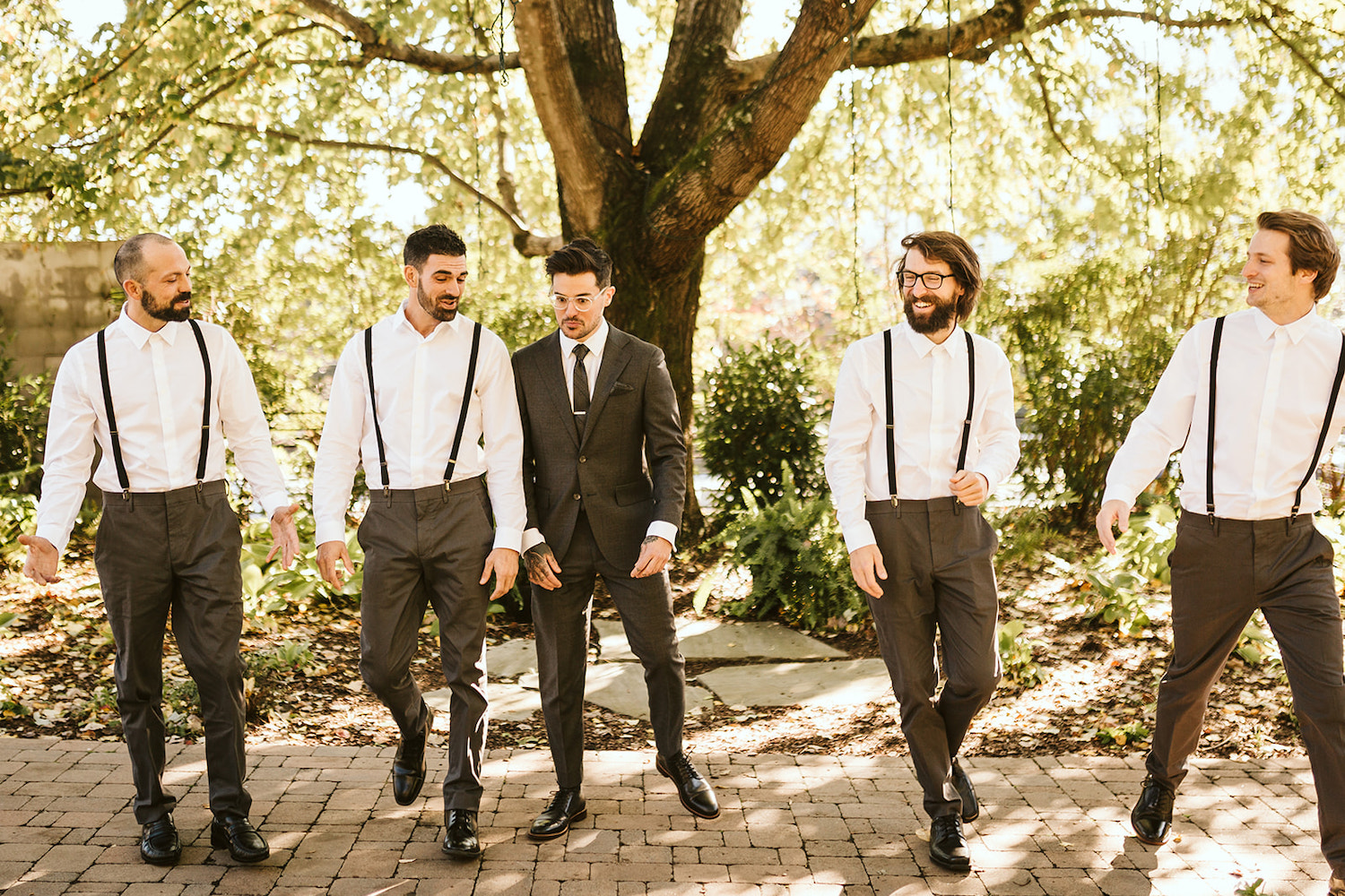 groom and groomsmen walk on a stone paver path under large tree