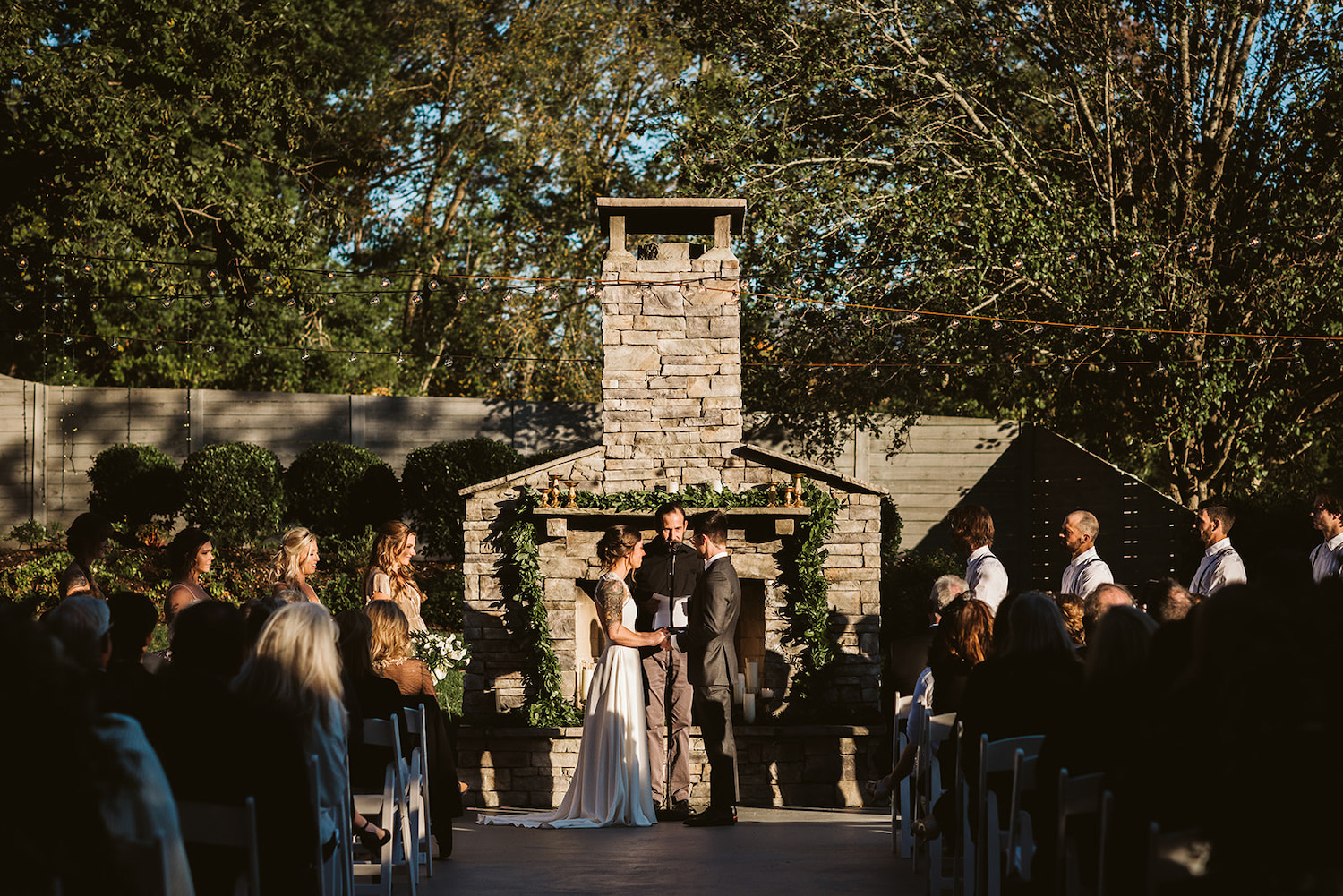 bride and groom stand with officiant in front of an outside stone fireplace