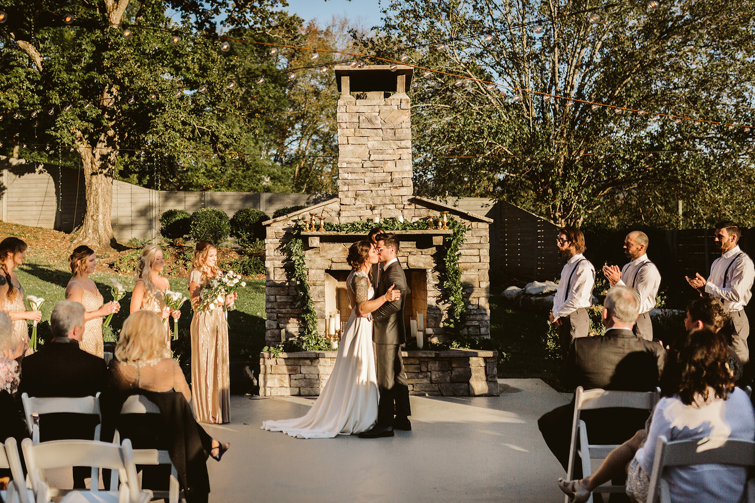 bride and groom share first kiss in front of an outside stone fireplace while wedding guests watch