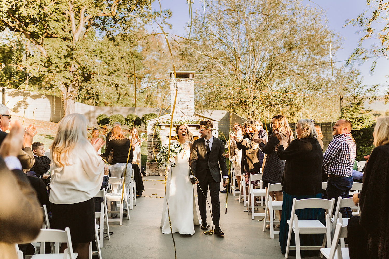 bride and groom walk down the aisle under gold streamers while guests clap