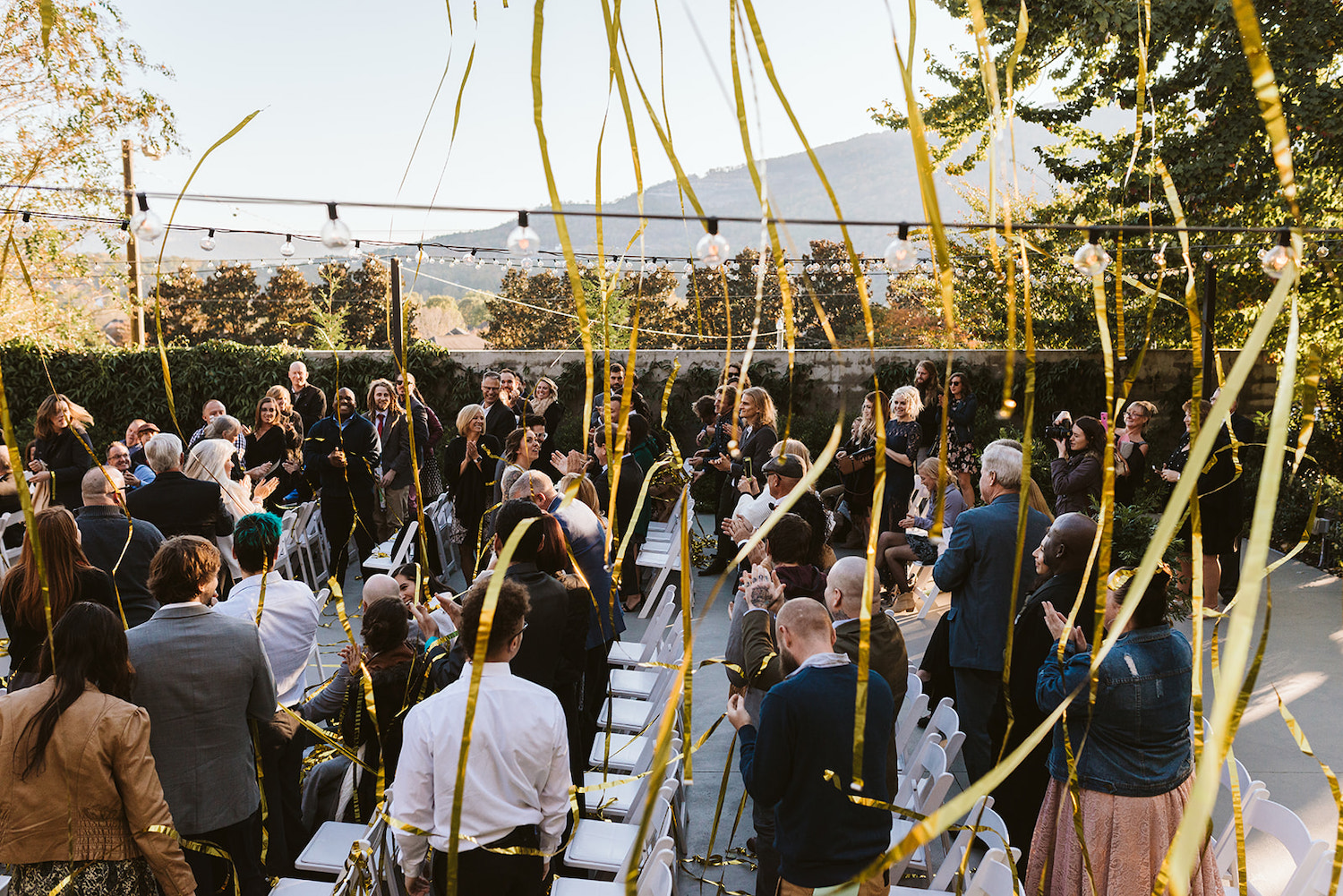 bride and groom walk down the aisle under gold streamers while guests clap