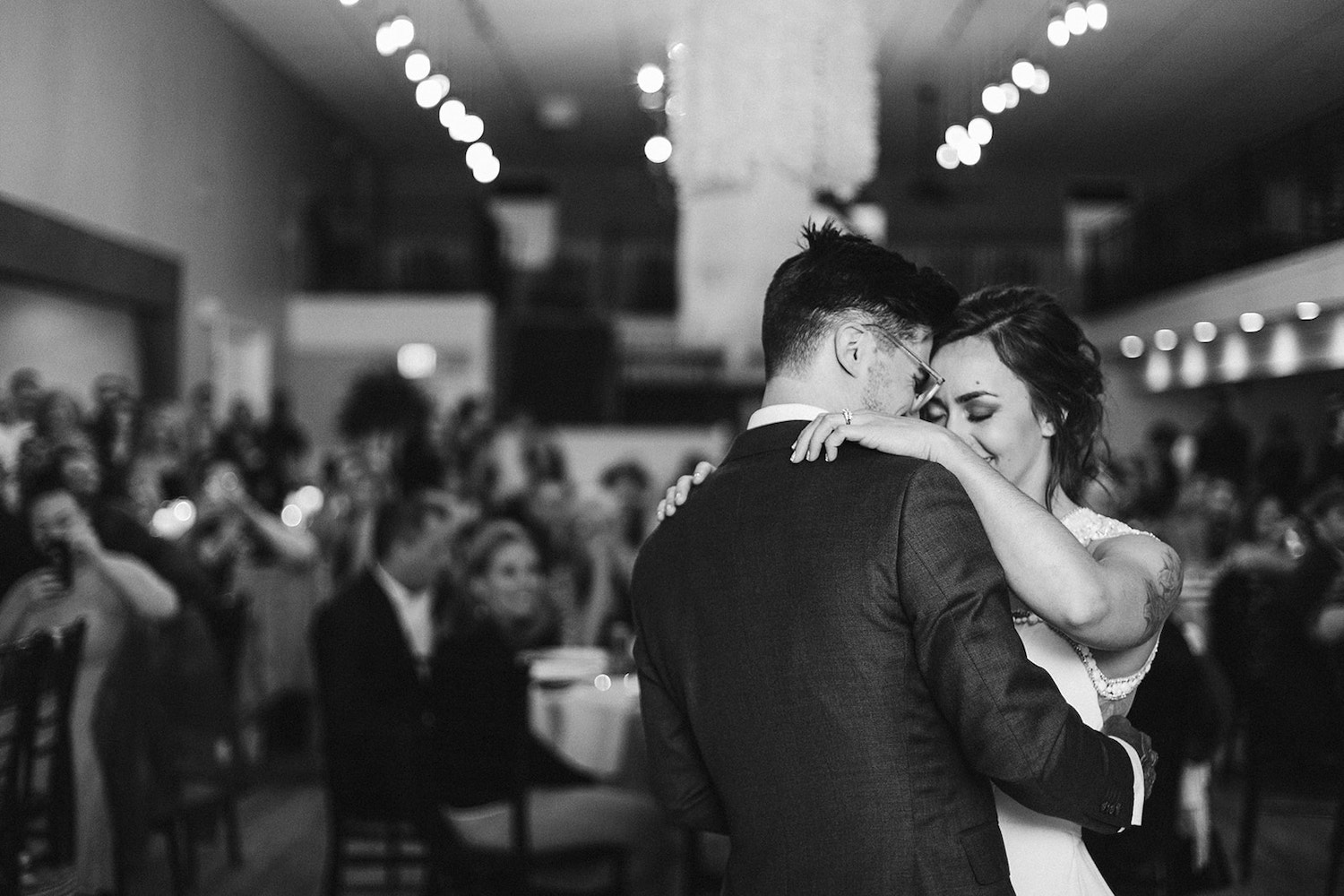 bride and groom smile at each other as they hold each other closely during their first dance while wedding guests watch