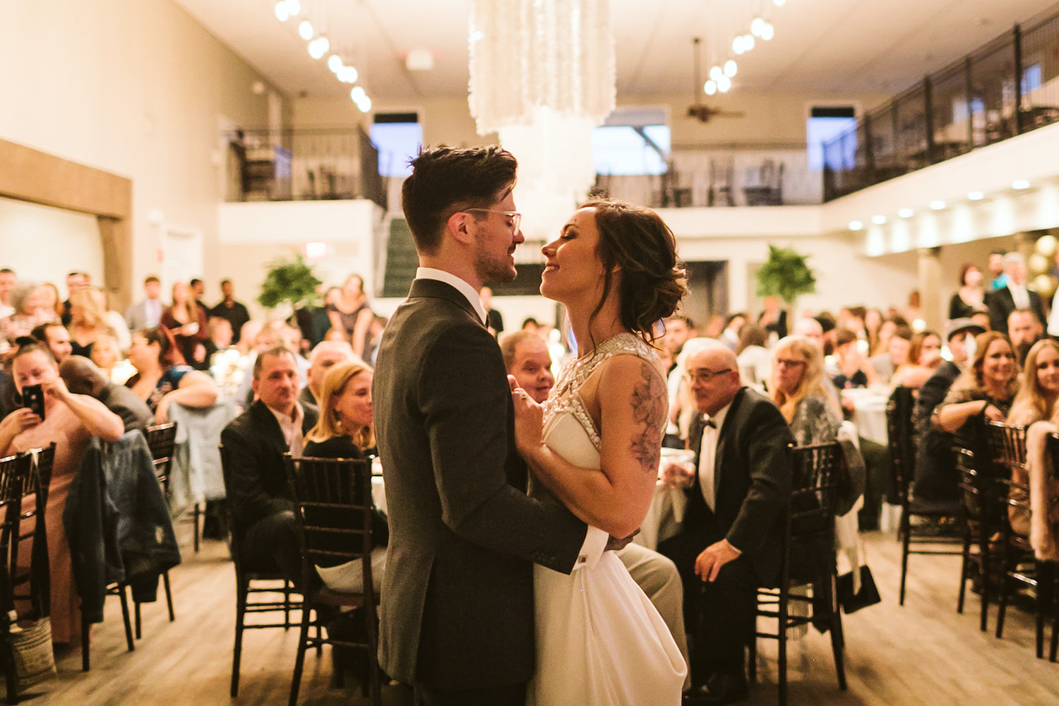 bride and groom smile at each other as they hold each other closely during their first dance while wedding guests watch