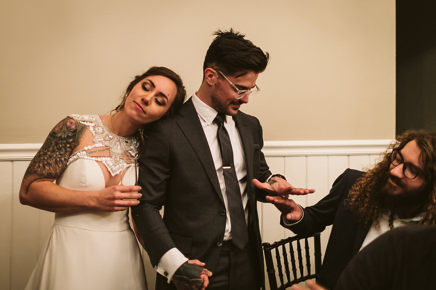 bride rests her head on groom's shoulder as he talks with a friend