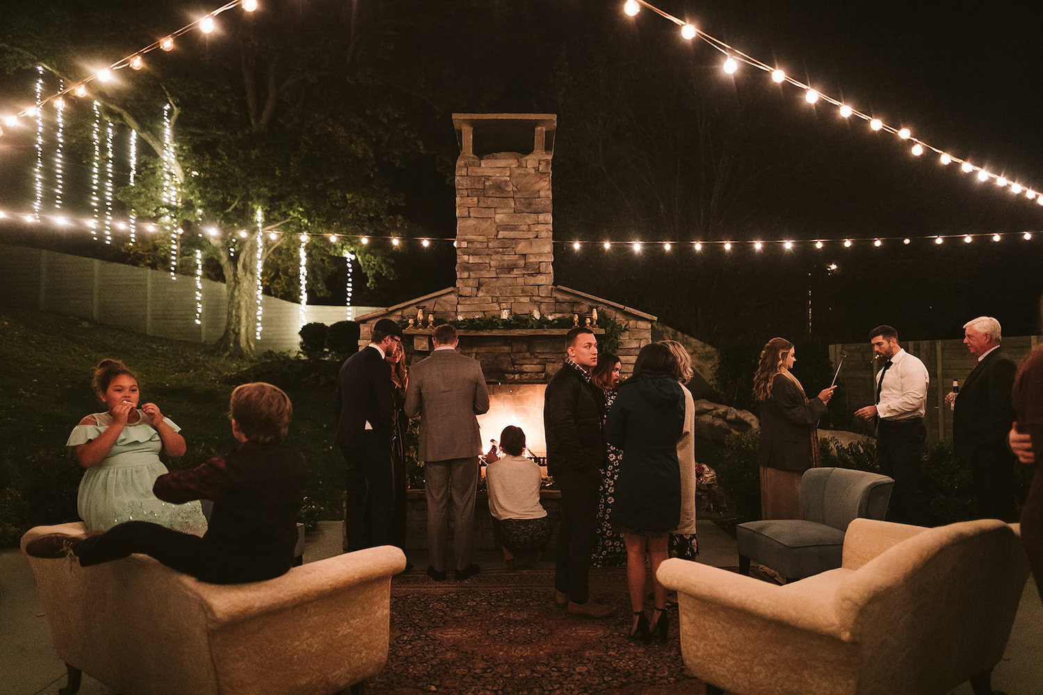 wedding guests gather near an outdoor stone fireplace under string lights