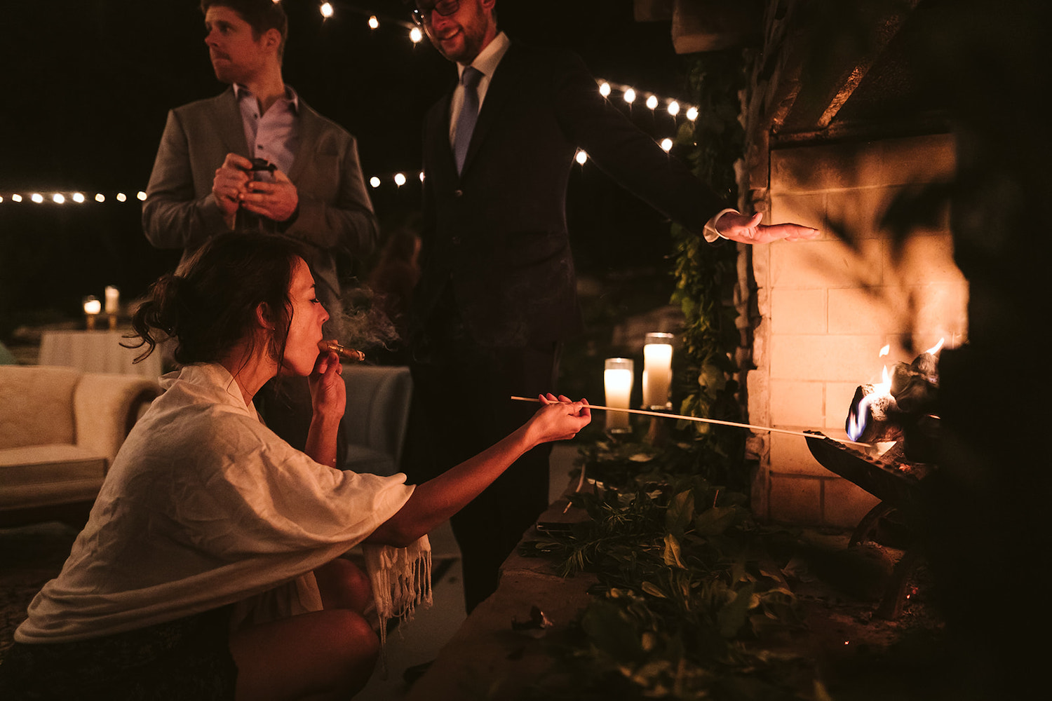 woman holds a cigar in one hand while she crouches in front of an outdoor fireplace roasting a marshmallow