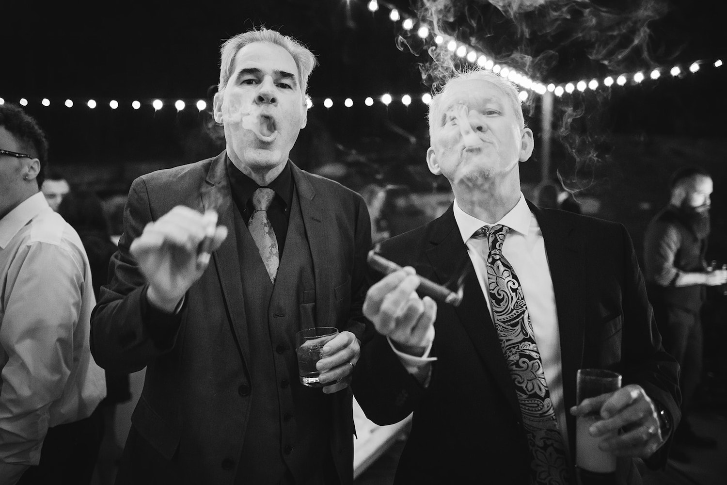 two older men in suits exhale puffs of cigar smoke in the night sky