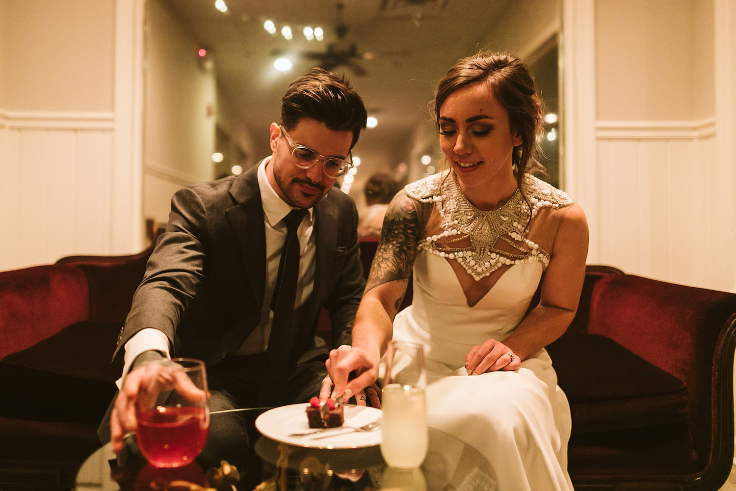 bride and groom sit close to each other on a low sofa. he reaches for cocktail and she cuts a small bit of cake with a fork