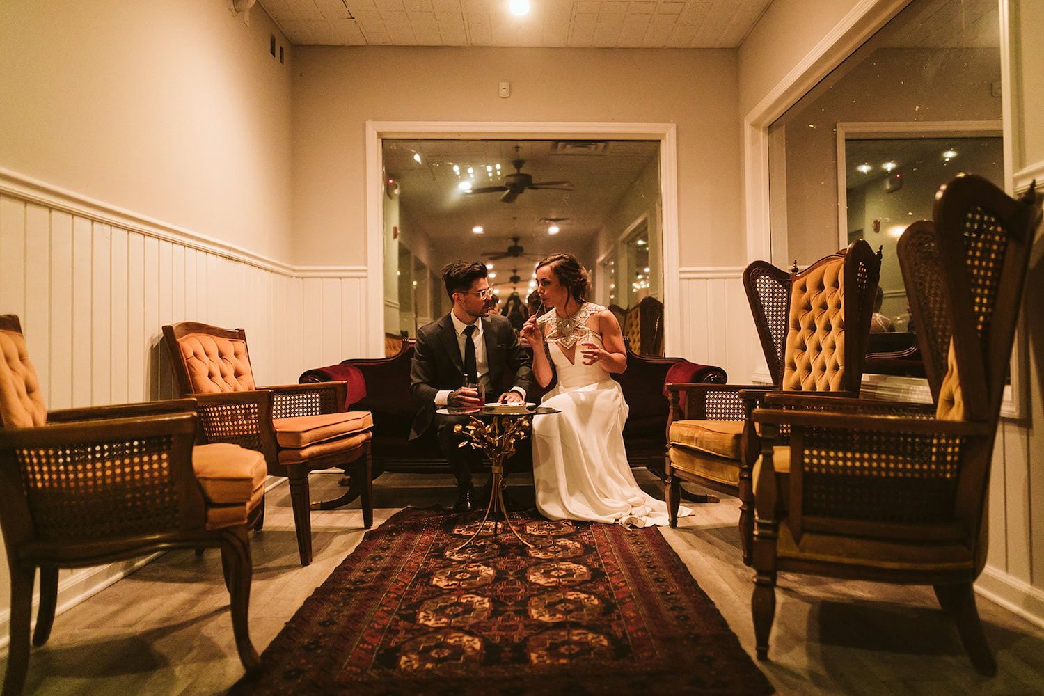 bride and groom sit together on low sofa enjoying cocktails and dessert