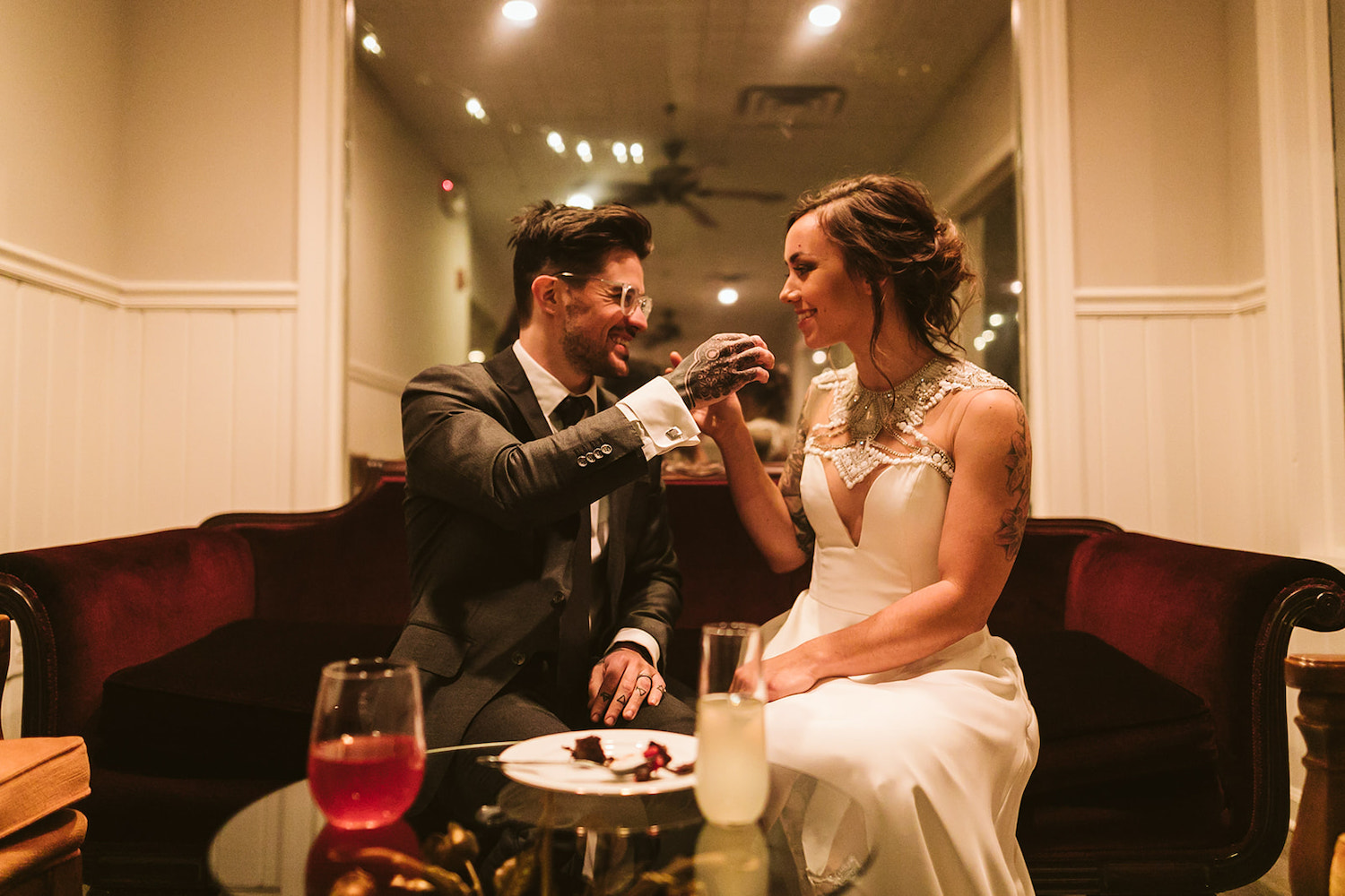 bride and groom sit on low sofa with hands outstretched to feed each other