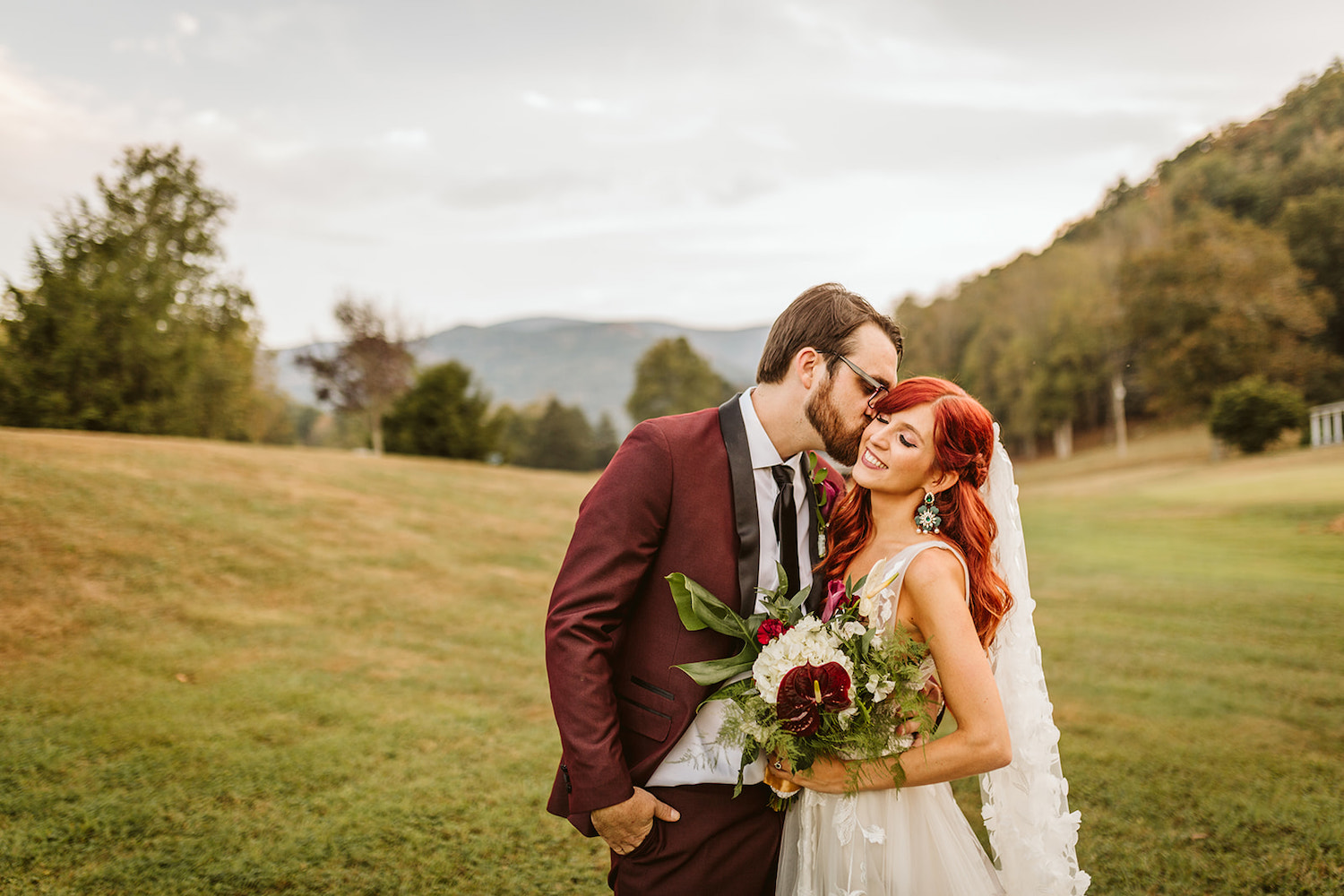groom in maroon suit kisses brides cheek in grassy field with hills in the background at their Roan Mountain wedding