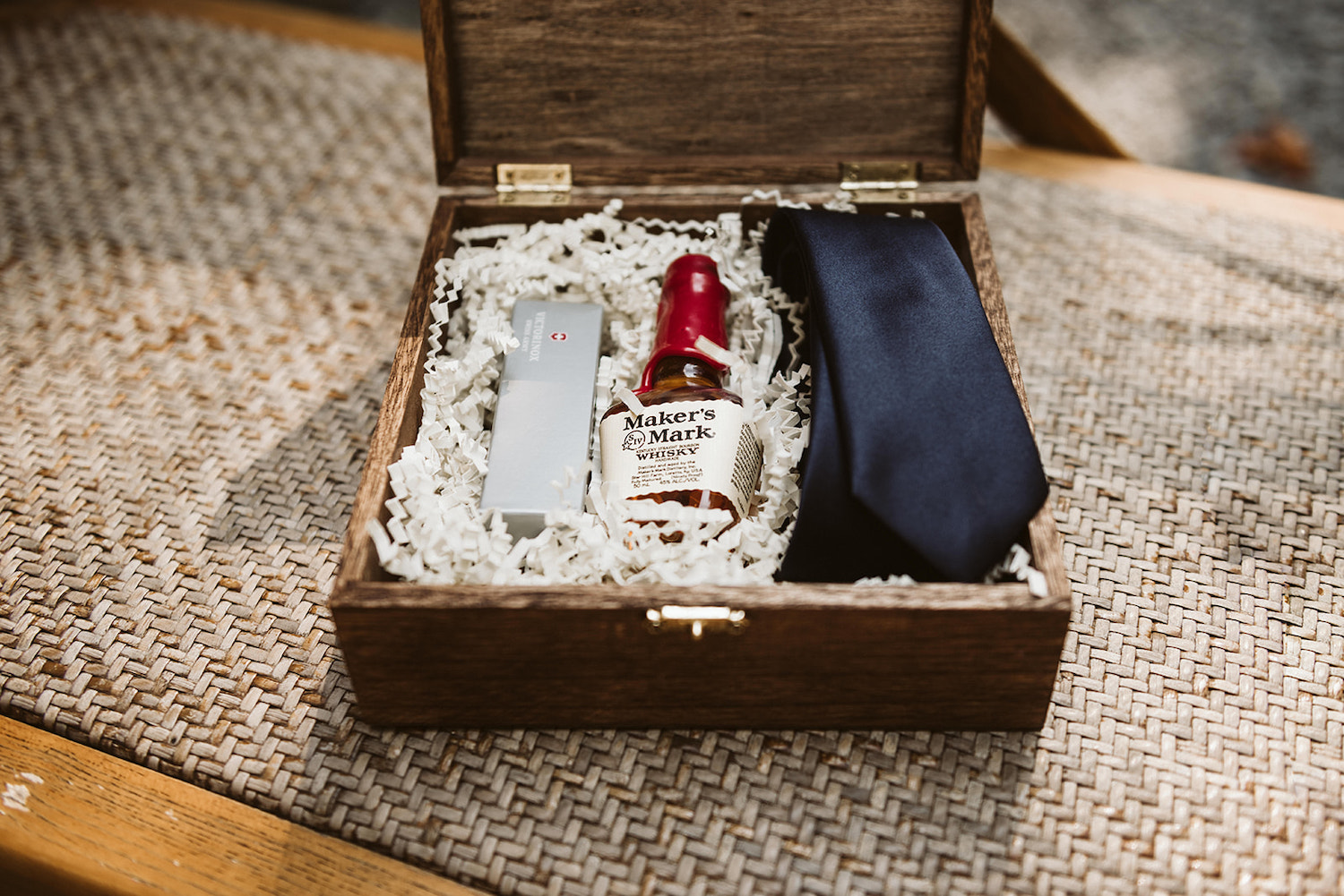 wooden box with lid hinged open contains a dark tie and small bottle of Makers Mark whisky