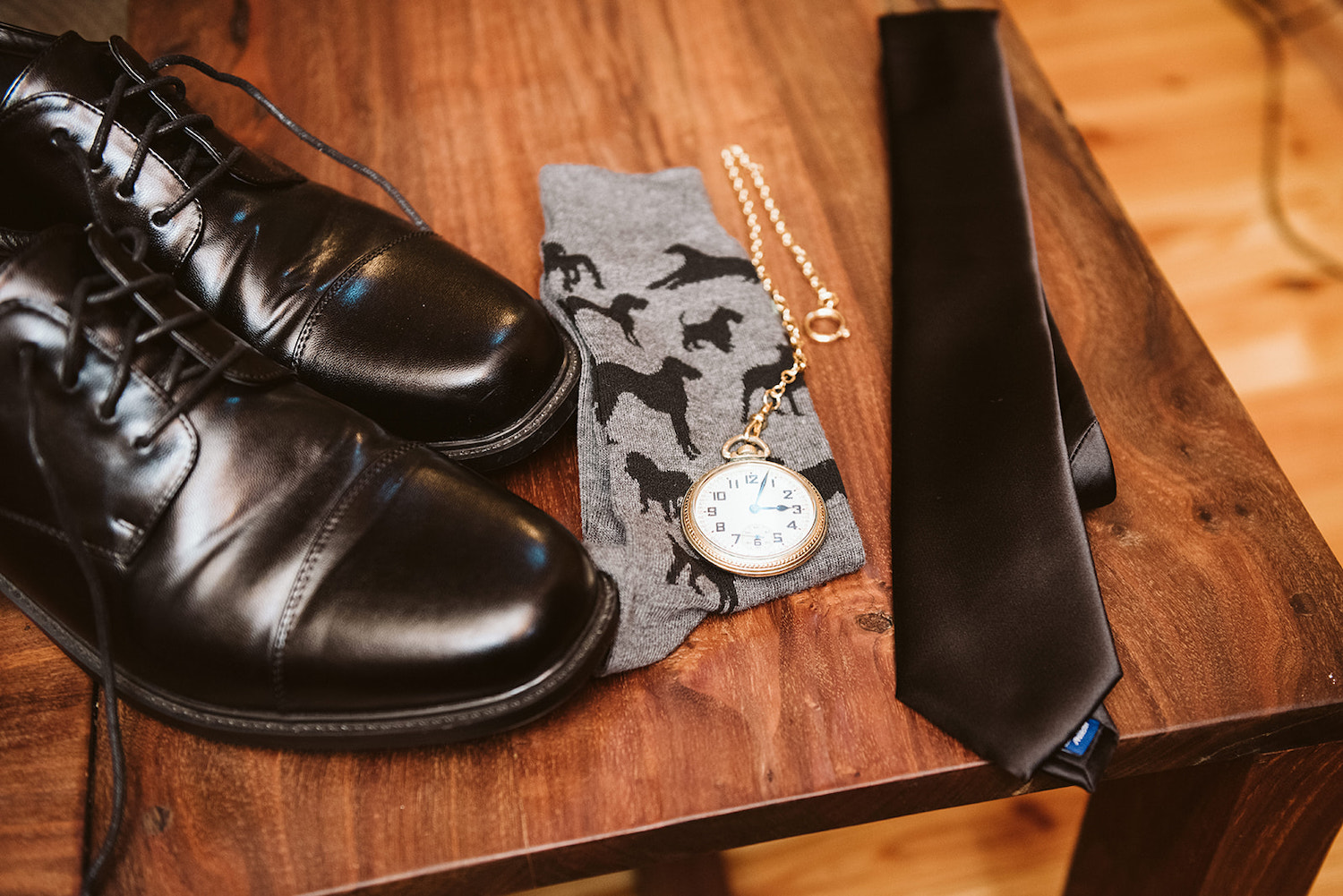 pocket watch sits on top of gray socks stitched with black dogs. Black mens shoes and black tie nearby.