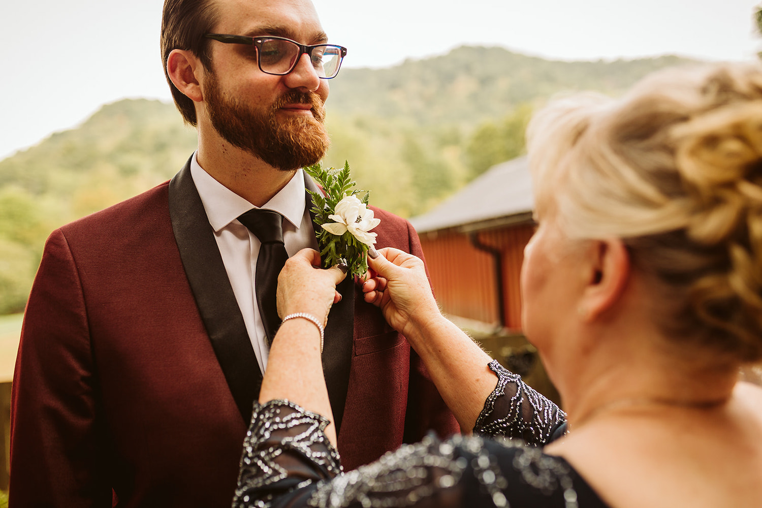 groom's mother pins a simple boutonniere of greens and white flower onto the black lapel of his maroon suit
