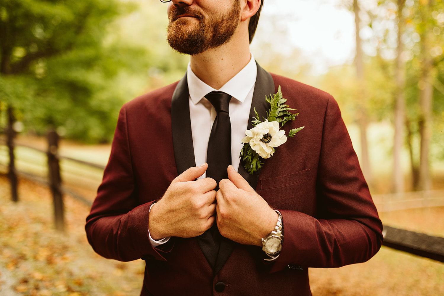 man grasps the black lapels of his maroon suit. a simple boutonniere of greens and white flower is pinned to his lapel.