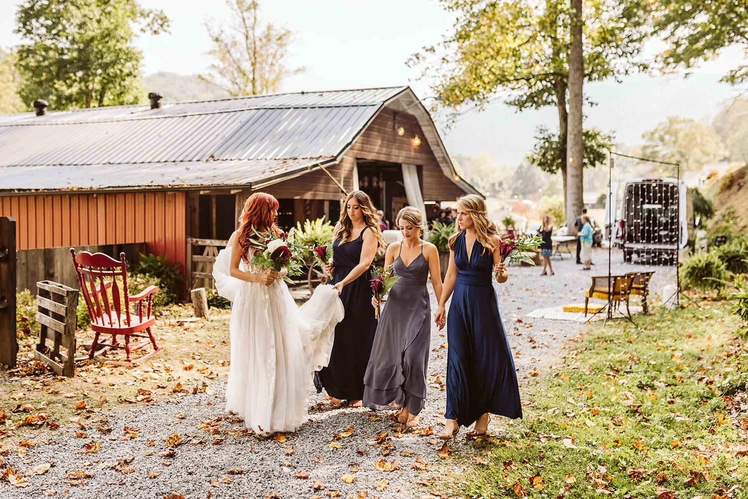 bridesmaids in long blue satin dresses walk with bride on a gravel path at her Roan Mountain wedding. one woman carries bride's dress train.