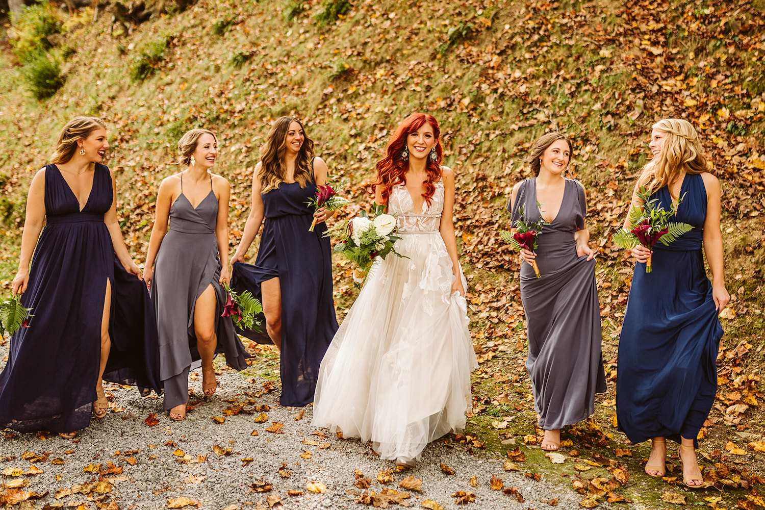 bride and bridesmaids in various blue satin gowns walk while holding their long gowns above the grass
