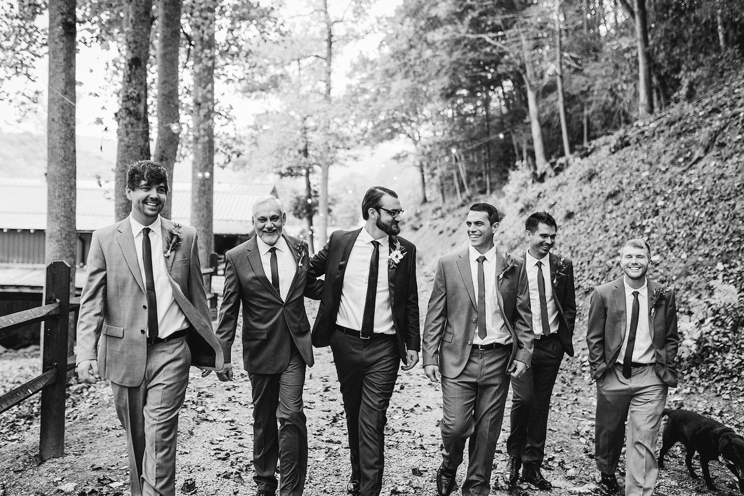 Groom in maroon suit and groomsmen in gray suits walk down a gravel path under tall trees
