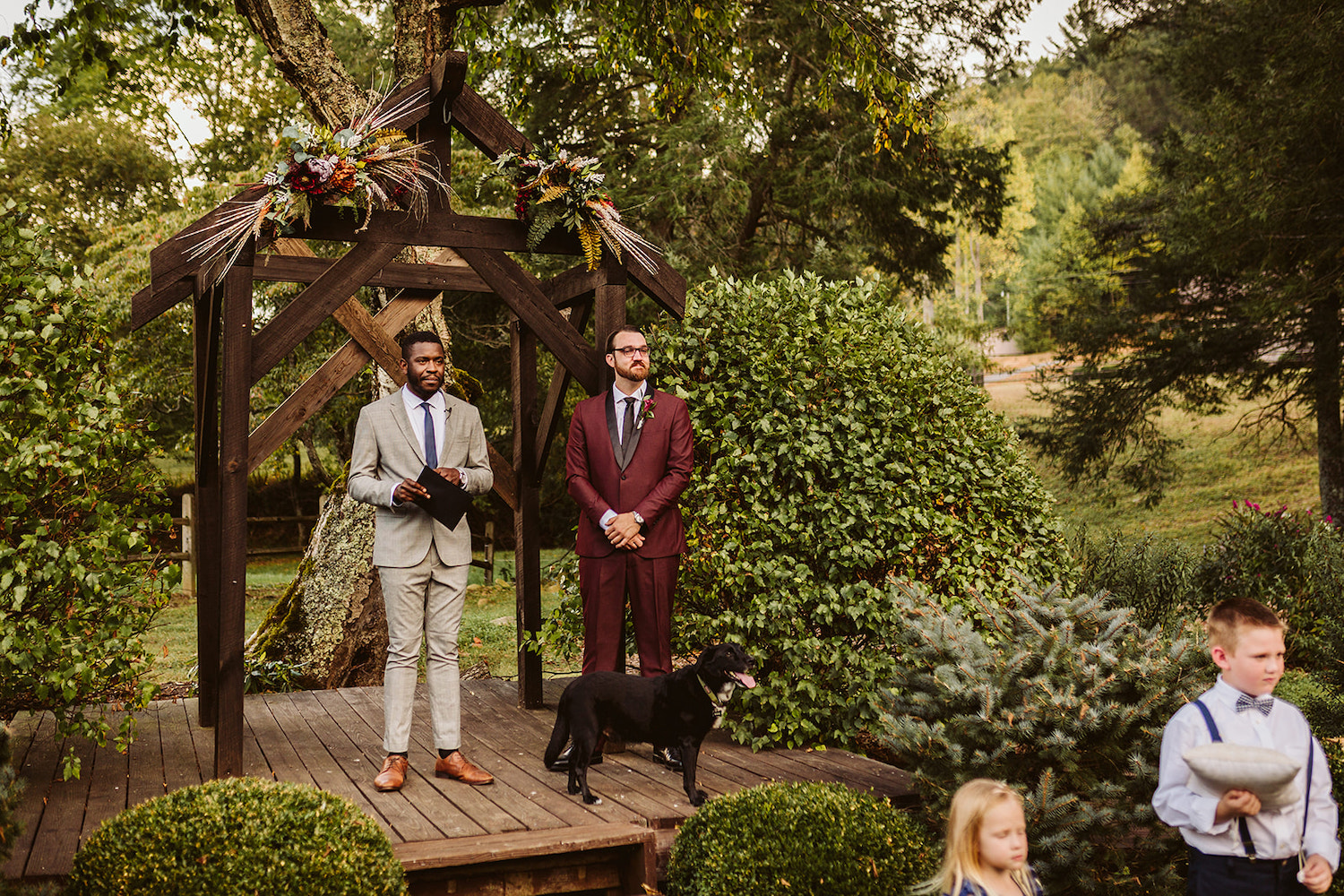 groom in maroon suit and officiant in tan suit stand on a wooden deck in front of a wooden archway