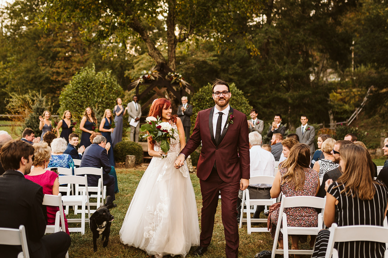 bride and groom walk down the aisle together with their black lab while guests clap for them