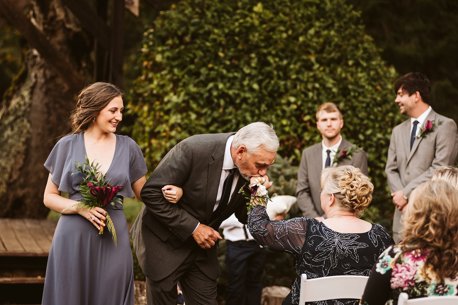 grooms father kisses grooms mother on the hand as he escorts a bridesmaid down the aisle